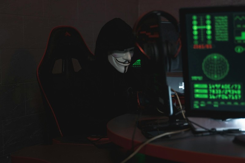 A hacker wearing a Guy Fawkes mask works at a computer in a dimly lit room