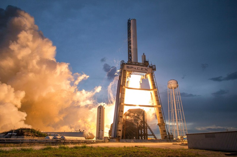 A SpaceX rocket on the launch pad with bright exhaust plumes against a dramatic dusk sky