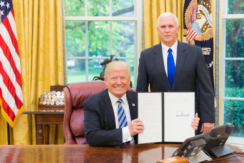 President Donald Trump signs a document at his desk in the Oval Office, with Vice President Mike Pence standing behind him.