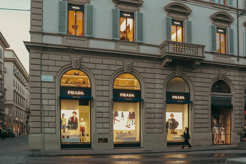 A Prada boutique storefront on a historic Florence street at dusk, with arched windows displaying mannequins and navy awnings bearing the Prada logo