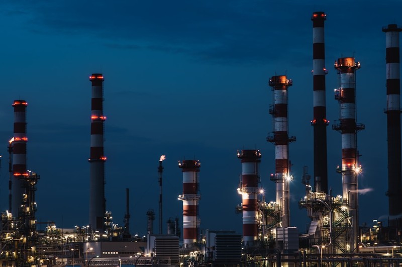 Oil refinery at dusk with illuminated smokestacks and industrial piping against a darkening sky