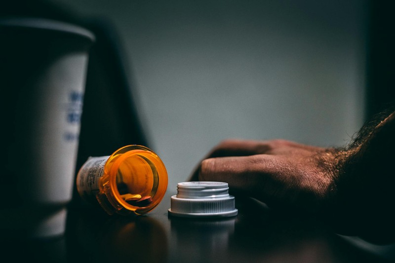 A prescription pill bottle lying open on a dark surface with a hand nearby, moody lighting