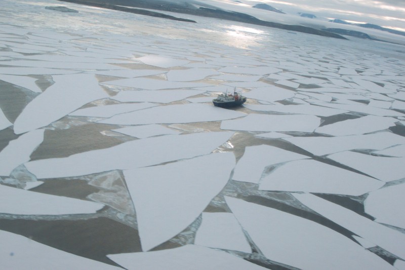 A small vessel navigates through fragmented sea ice under an overcast sky in a vast frozen sound.