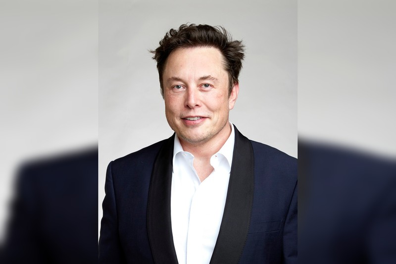 Elon Musk poses for a portrait wearing a dark suit and white shirt against a neutral gray background
