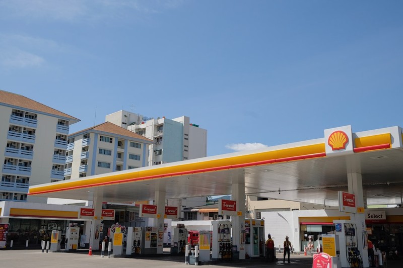 A Shell gas station with fuel pumps under a yellow and red canopy against a clear blue sky in an urban setting