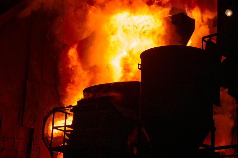 A dramatic blast furnace blazing with intense orange and yellow flames inside a dark steelworks, with industrial machinery silhouetted against the fire.
