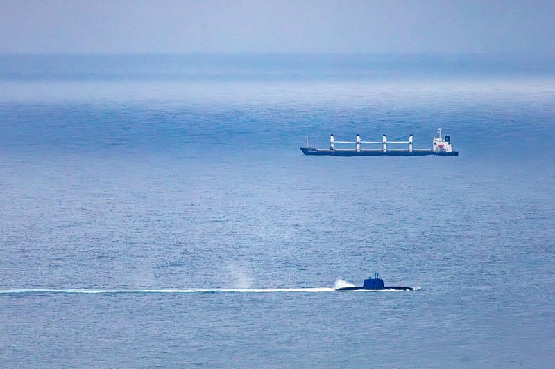 A submarine and a cargo ship navigate the open ocean under an overcast sky