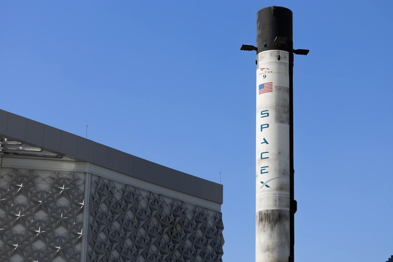 A SpaceX Falcon 9 rocket displayed vertically outdoors against a clear blue sky with the SpaceX logo visible on its body.